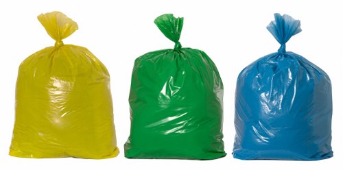 The image displays three large, tightly filled rubbish bags made of glossy plastic in yellow, green, and blue colors, arranged in a row on a paved outdoor surface. The yellow bag is positioned on the left, the green in the middle, and the blue on the right, each with a gathered and tied top. The bags appear to contain waste materials, possibly household or commercial rubbish, and are standing upright with slight creases and reflections of light on their surfaces. The background is minimal and out of focus, suggesting an outdoor area such as a driveway or pavement adjacent to a property in Golders Green, with no other visible objects or environmental features. The arrangement and material of the bags are typical of waste collection or rubbish removal services provided by companies like Commercial Waste Removal Golders Green, which handle general rubbish disposal in the local area.