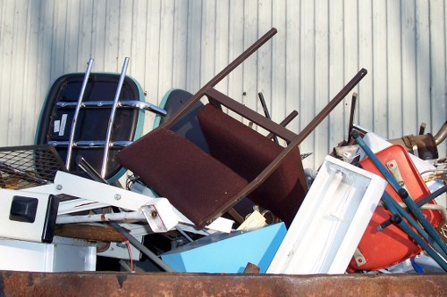 A large pile of mixed household and garden rubbish stacked outdoors against a corrugated metal wall, including items such as broken wooden chairs with dark brown upholstery, a white plastic drawer unit, a red plastic container, a metal wire basket, a black plastic chair frame, and other discarded furniture and fixtures. The debris is piled haphazardly, with some objects leaning or partially obscured, indicating a typical scene of waste awaiting collection or disposal at a commercial waste removal site near Golders Green. The surrounding environment suggests an open area used for waste storage, with natural daylight illuminating the scene. Commercial Waste Removal Golders Green provides rubbish clearance services in this area, supporting local businesses and residents in managing bulky and unwanted waste efficiently.