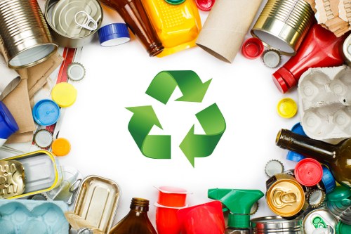 A variety of recyclable waste materials arranged in a circular pattern around a large green recycling symbol at the centre. The waste includes aluminium cans with metallic finishes in silver, gold, and green, plastic bottles in red, yellow, and clear with caps intact, cardboard tubes and sheets in brown, and plastic containers in various colours such as blue, red, and green. The background is white, highlighting the different textures and colours of the waste items, which are placed on a flat surface. This image signifies the importance of rubbish recycling and waste separation, relevant to services like those offered by Commercial Waste Removal Golders Green, operating within the NW11 postcode area, supporting local waste management needs in the Golders Green district.