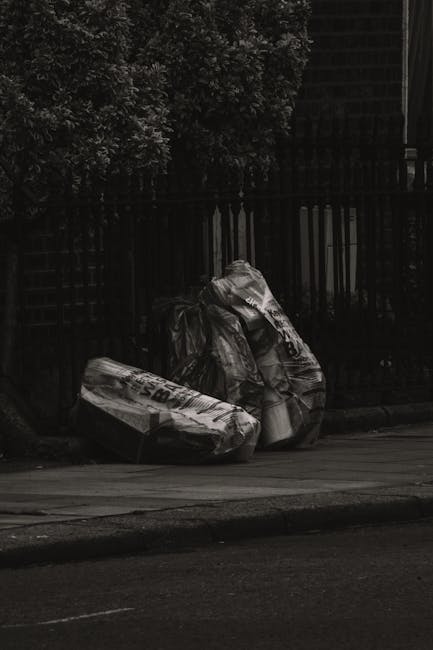 Two large black rubbish bags, made of thick plastic with a glossy finish, are placed on the curbside in front of a black metal fence. One bag appears bulging and tied at the top, indicating it is fill