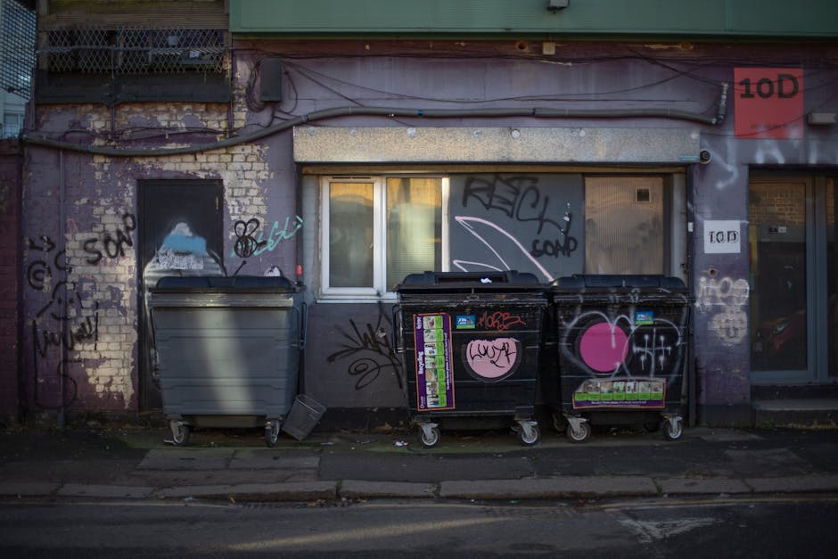Two large black rubbish bags, made of thick plastic with a glossy finish, are placed on the curbside in front of a black metal fence. One bag appears bulging and tied at the top, indicating it is fill